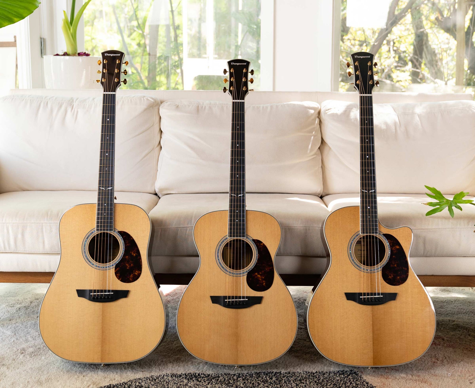 Three Orangewood Guitars leaned against a white couch with plants in the background