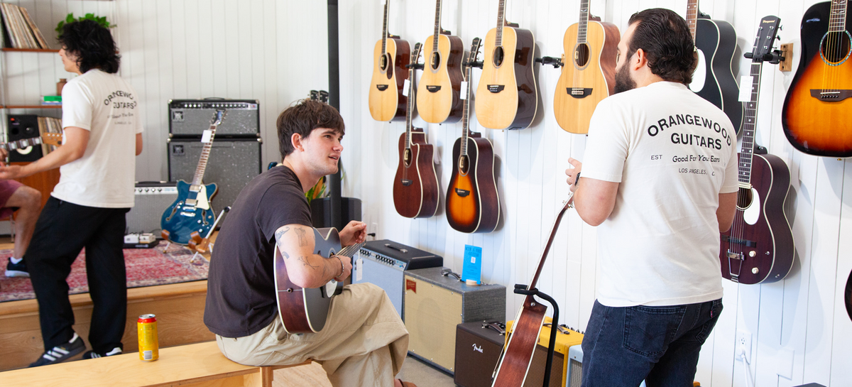 Three men in a guitar shop or studio, with two wearing white t-shirts that read 'ORANGEWOOD GUITARS EST. 2012' and one holding an acoustic guitar.