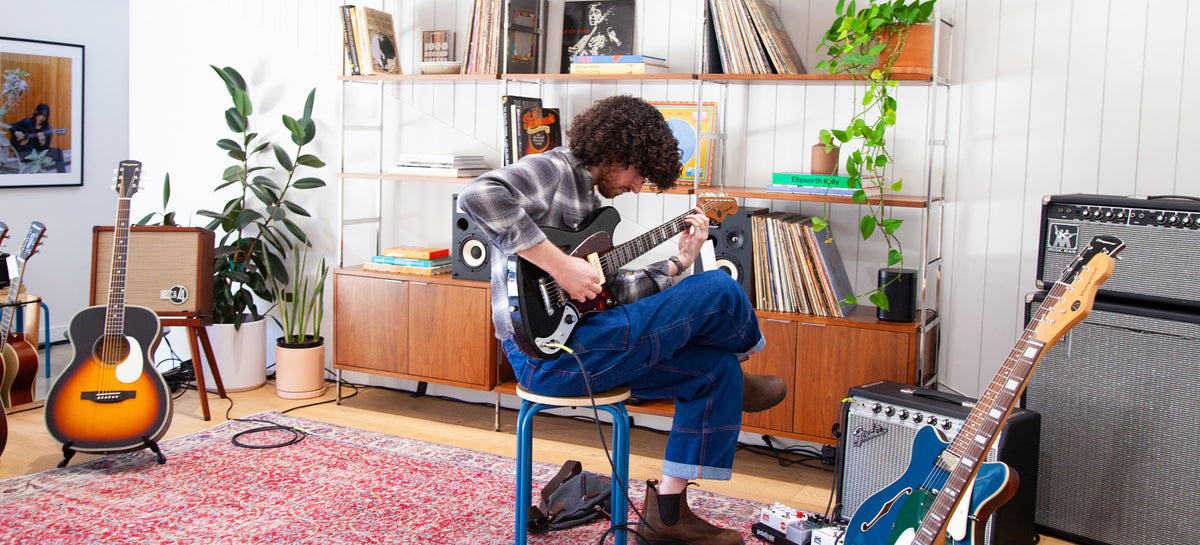A person with curly hair sits on a blue stool in a music room, playing an electric guitar. An acoustic guitar stands on a stand to the left, and another blue electric guitar leans against a speaker on the right.