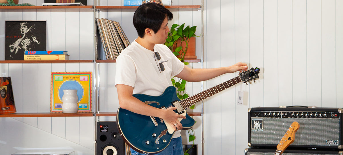 A man in a white t-shirt stands in a room holding a blue electric guitar, with a guitar amplifier and a microphone visible in the background.