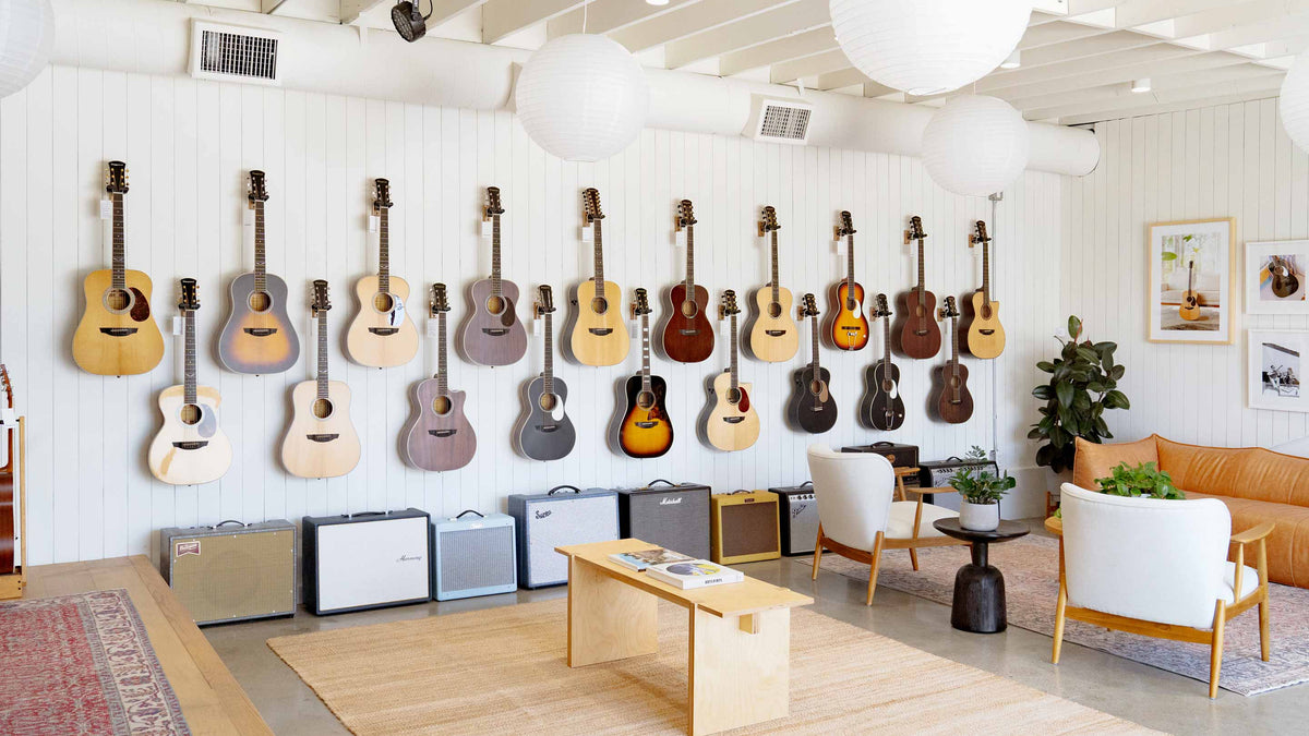 A large collection of acoustic and electric guitars displayed on a white paneled wall in a bright, modern showroom. Several guitar amplifiers sit on the floor below, and a small seating area with white chairs, a wooden table, and a tan sofa is visible in the foreground and to the right.