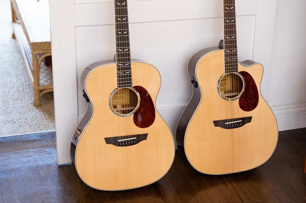 Two spruce Orangewood Melrose guitars with pick guards lean against a white door 