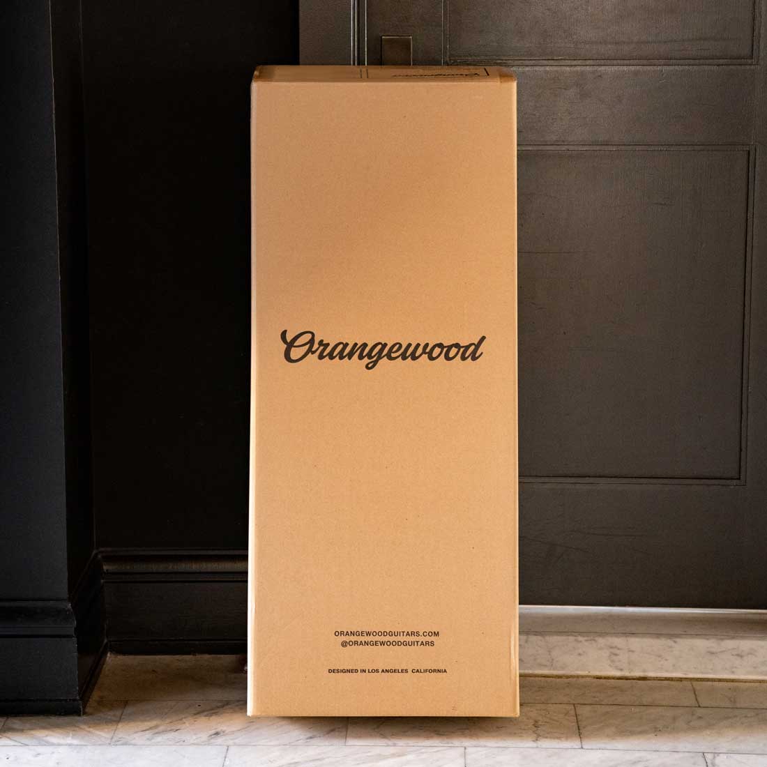 An Orangewood shipping box sits in front of a black front door on marble floors