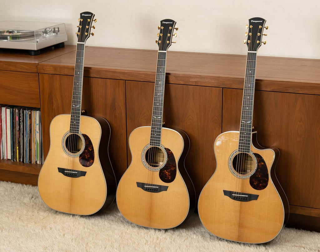 Three Orangewood torrefied spruce Topanga guitars with pick guards lean against a wood cabinet